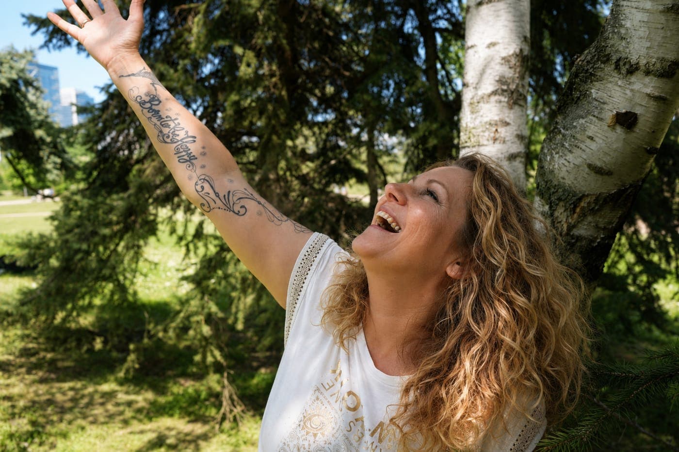 Christine Gaubert, praticienne LaHoChi, riant, un bras tendu vers un arbre dans un parc ensoleillé